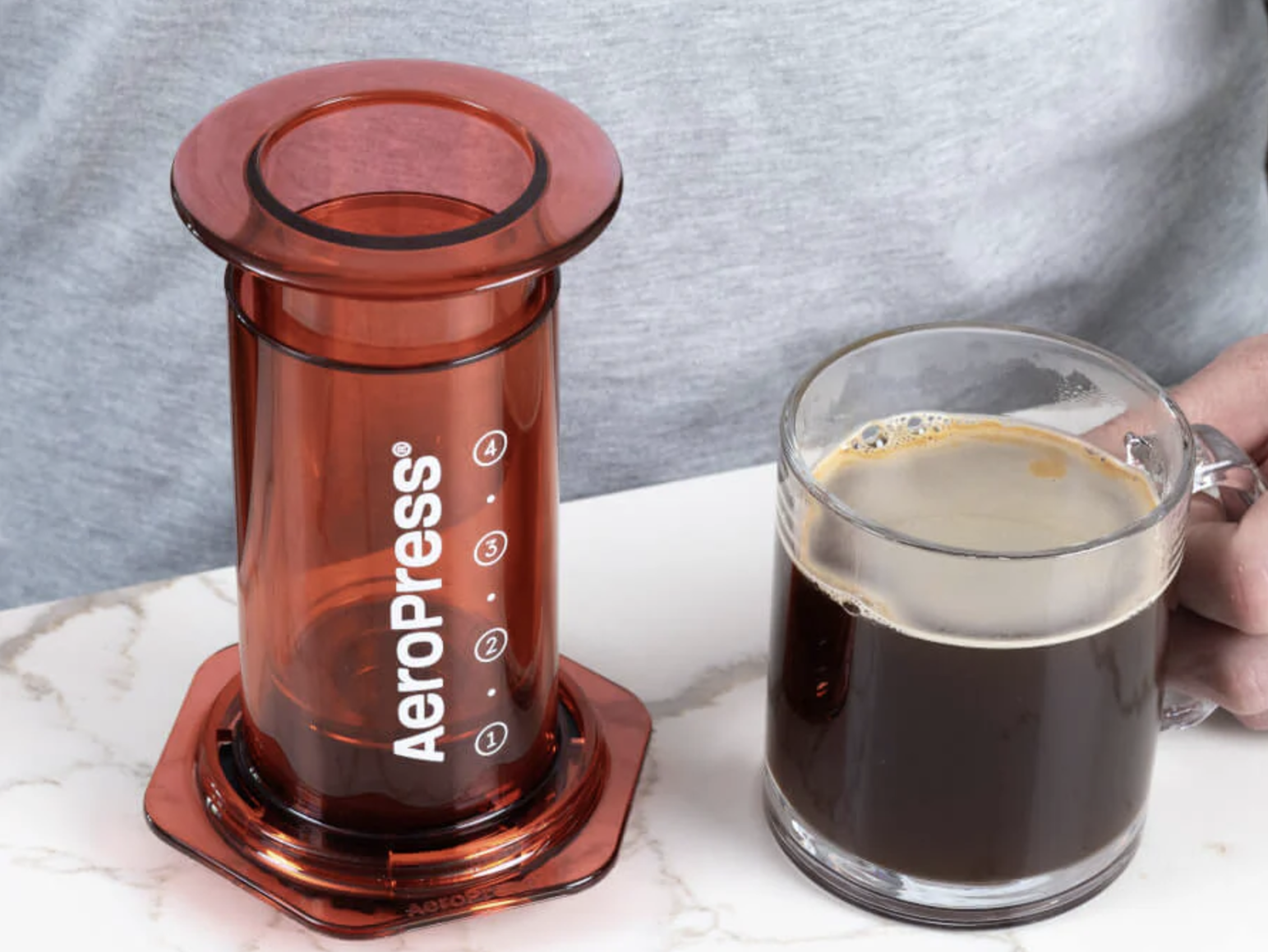 A person holds an AeroPress and a cup of coffee.